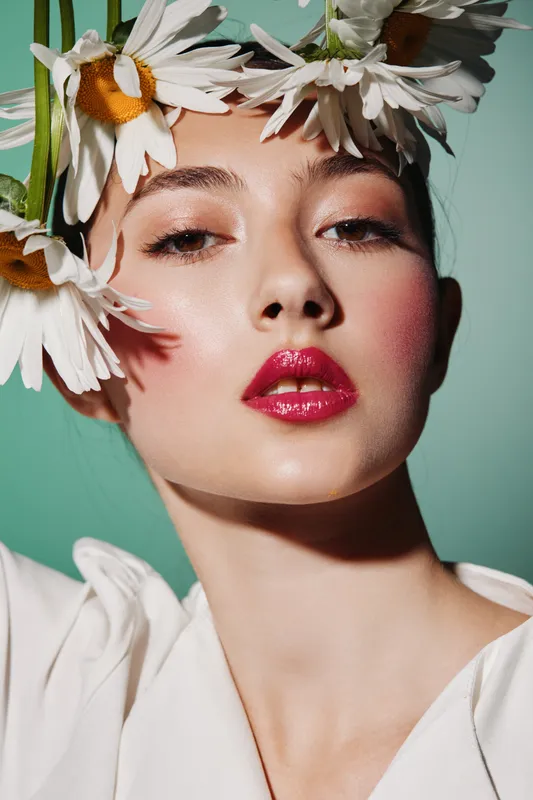 Close-up portrait of a creative female beauty model with vivid glossy lipstick and white floral crown on pale skin, photographed against mint background in fashion concept for beauty editorial.