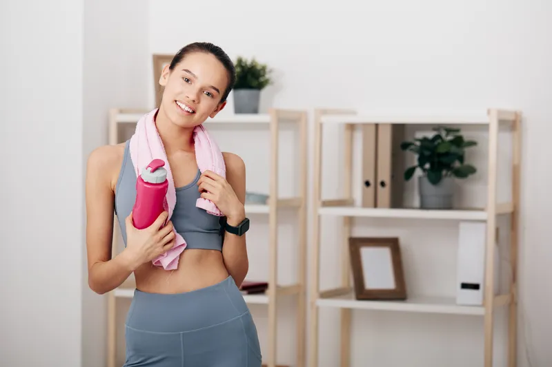 Active woman in sportswear with towel draped around neck and a water bottle, standing in a neat home gym setup ready for a workout, promoting fitness, health, and daily training.