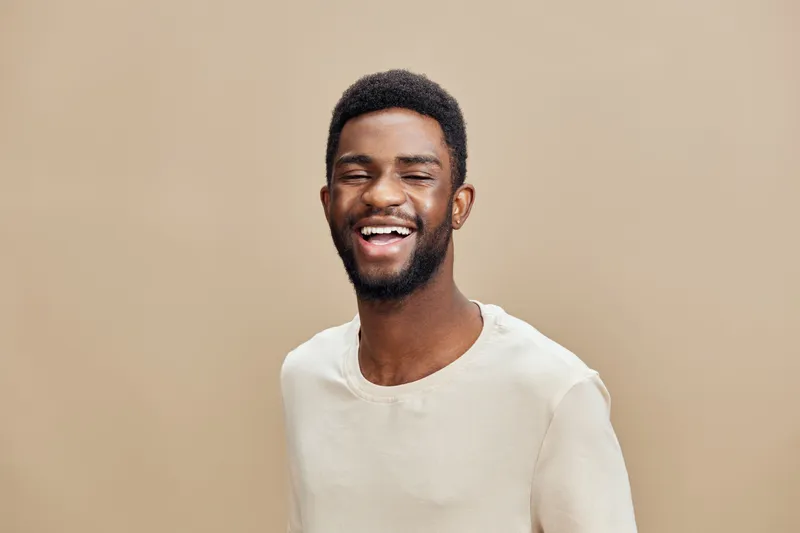 Smiling young Black man in a light beige shirt, expressing happiness against a soft beige background, perfect for concepts of joy and positivity