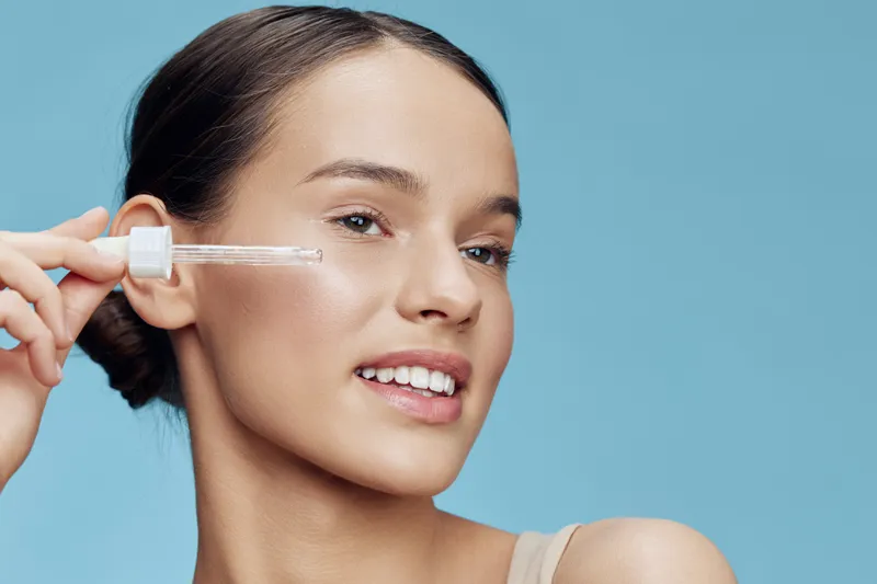 Young woman applying serum on her face, glowing skin, smiling expression, blue background, healthy beauty routine, skincare essentials