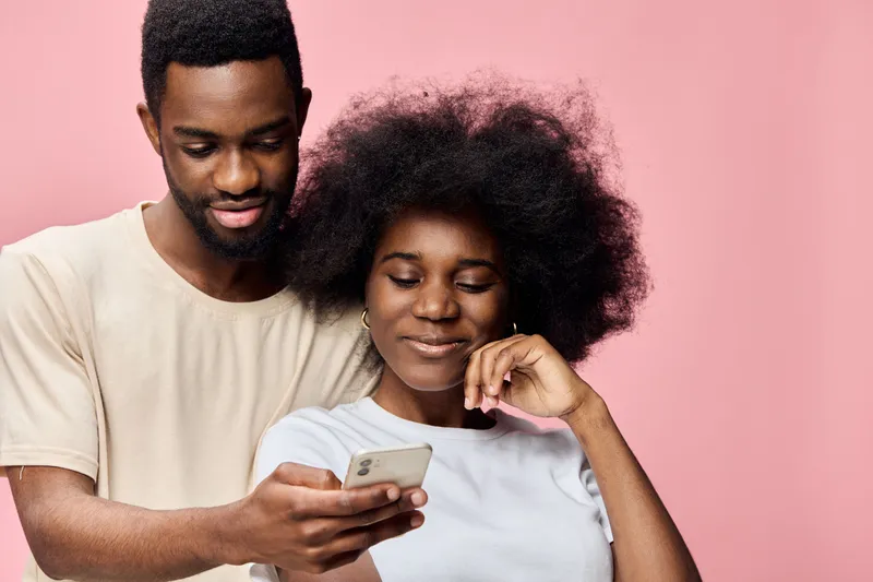 Young African couple sharing a joyful moment while looking at a smartphone against a pink background, showcasing happiness and love in a vibrant, modern setting