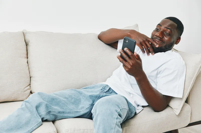 Relaxed young African American man enjoying content on his smartphone while lounging on a light colored sofa at home