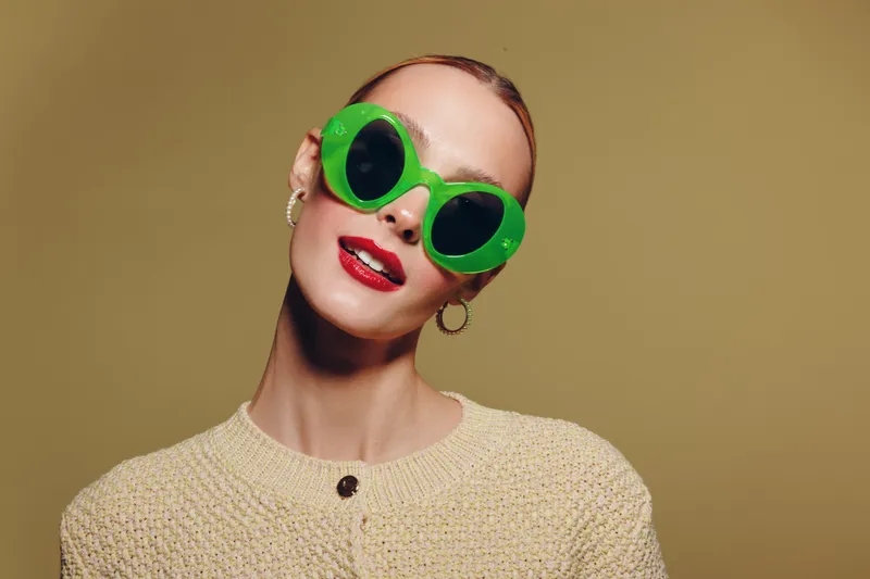 Young woman with slicked hair wears oversized green sunglasses and red lipstick, posing against beige background. Fashion concept with bold accessories and confident expression.
