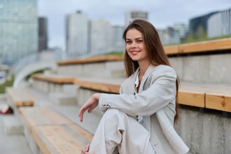 Confident smiling woman in light business suit sitting outdoors on urban stairs under cloudy sky