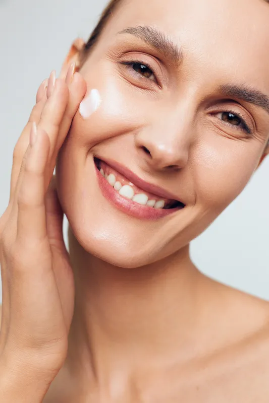 Beauty skincare woman applies cream to her face, smiling portrait, showcasing moisturizer use in a bright studio, healthy glow and clear skin, natural expression.