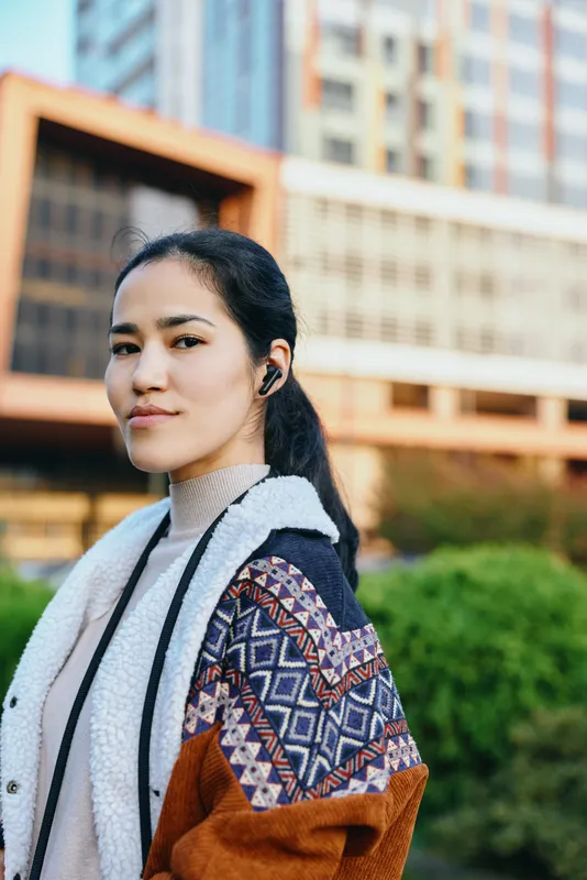 Woman wearing a patterned shawl and casual coat in an outdoor urban setting, listening to headphones, confident and stylish amid street and architectural backdrop