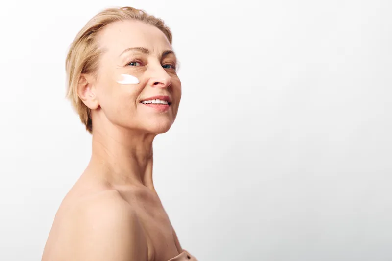 Smiling mature woman with short blonde hair applying moisturizing cream on her cheek. Close-up portrait on white background. Concept of skincare, aging beauty, and self-care routine.