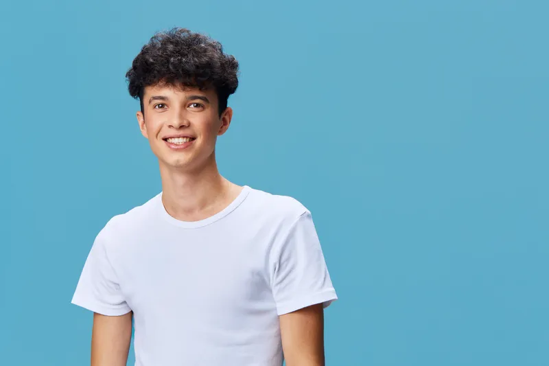 smiling young man in white t-shirt with curly hair standing against a blue background, showing positive energy and casual style