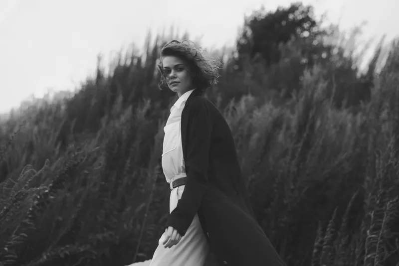 Woman walks through tall grass in a flowing coat, wind in hair, monochrome fashion shot with moody atmosphere, cinematic lighting, and subtle grain for dramatic visuals