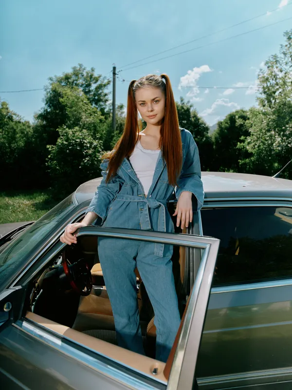 Young woman in denim jumpsuit standing by a vintage car, showcasing casual style, outdoors among green trees, with a bright and sunny background.