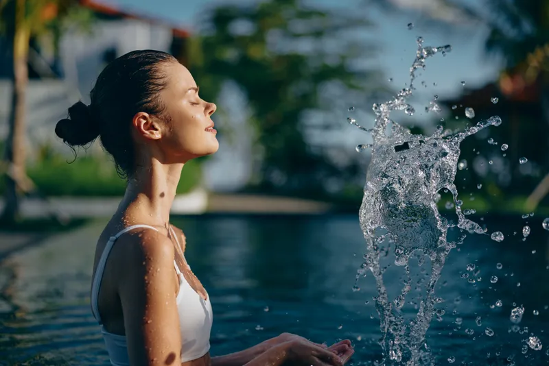 Young woman with a bun in a white tank top sits by a pool, water spray arcs in front, capturing a refreshing outdoor lifestyle moment of relaxation and sunshine.
