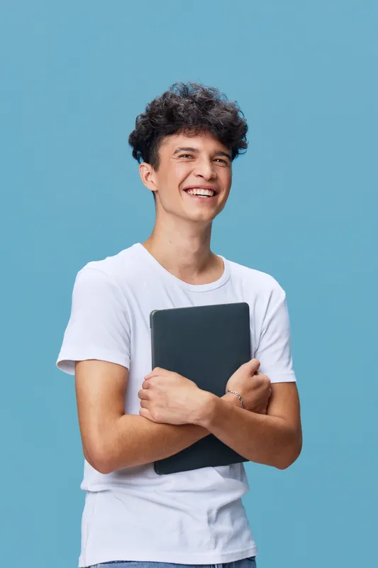 Smiling young man holding a laptop with arms crossed, standing against a blue background, wearing a white t-shirt, casual and cheerful expression.