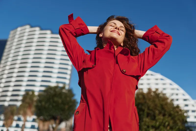 Smiling woman wearing a red coat enjoying sunny outdoor scene against modern buildings. Bright daylight highlights fashion, confidence, and a carefree mood in an urban setting.