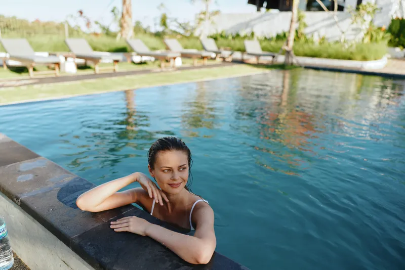 A relaxed woman in a white swimsuit enjoys leisure time in an outdoor pool surrounded by lounge chairs and greenery on a sunny day. Concept for summer lifestyle and wellness.
