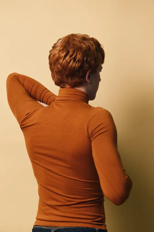 Redhead man standing with hand behind head wearing brown turtleneck sweater facing beige wall in casual outfit and relaxed pose for fashion or lifestyle portrait.