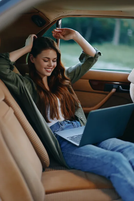 Happy young woman working on a laptop in a cozy car interior, enjoying her time while wearing casual clothes, featuring a relaxed vibe and natural background.