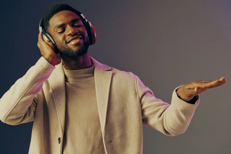 Young Black man enjoying music with headphones, smiling and swaying in a stylish outfit, conveying joy and relaxation in an artistic setting