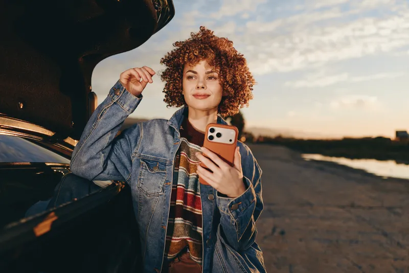 Woman lifestyle outdoors, curly hair smiling, holding smartphone near car, denim jacket with sleeves raised, warm sunset light, relaxed pose, natural colors, soft evening breeze, calm travel mood