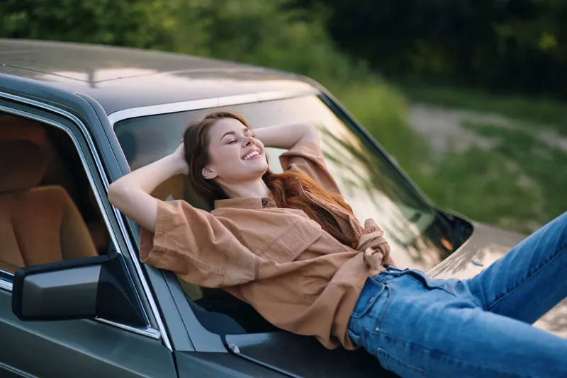 Young woman enjoying a sunny day outdoors, lounging on a vintage car hood, wearing casual fashion, showcasing happiness and relaxation during summer.