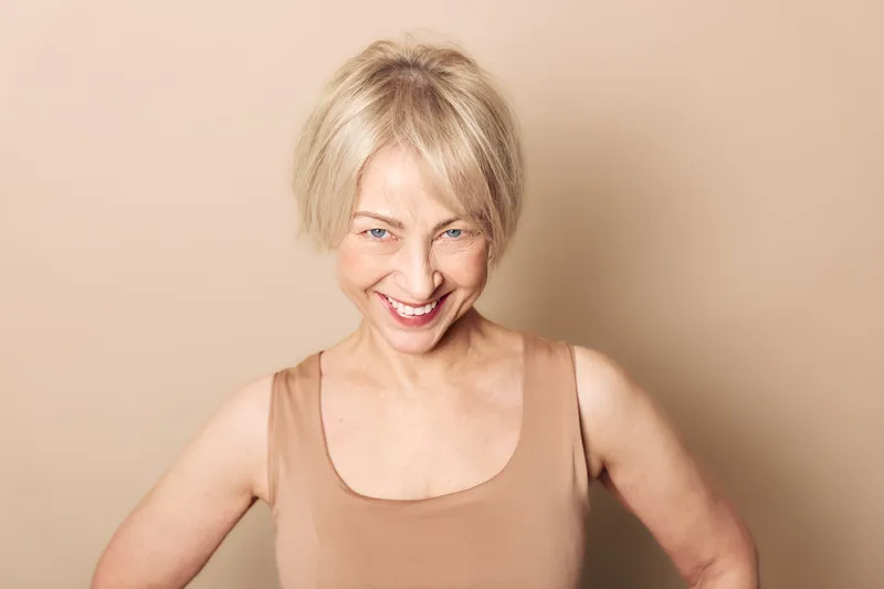 Smiling middle aged woman with short blonde hair wears beige tank top, looking confidently at camera on neutral background. Concept of happiness, confidence, mature beauty and wellness.