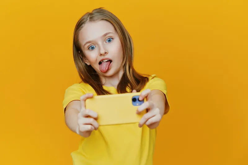 Playful girl taking a selfie with a yellow smartphone against a bright yellow background, showcasing joy and enthusiasm in a vibrant summer atmosphere