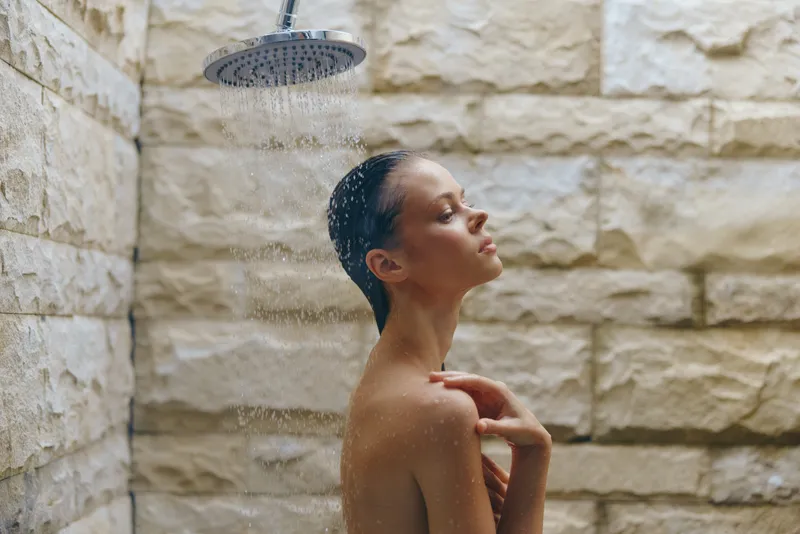 young woman enjoying refreshing outdoor shower under rainfall, relaxed pose, natural stone wall background, wet skin glistening in soft light