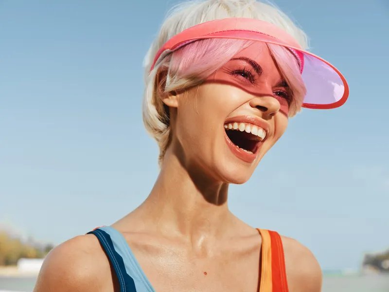 Vibrant portrait of a smiling woman at the beach wearing a pink visor, sunlit skin and a bright blue sky backdrop, capturing energetic summer vibes.