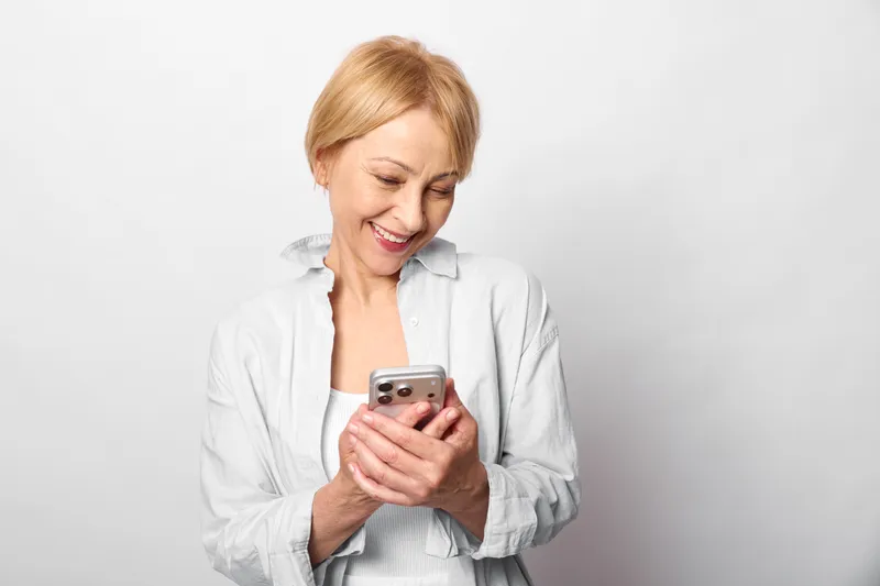 Woman smartphone smiling senior communication technology indoors white portrait casual, elderly woman in studio holds phone and reads messages, soft light, happy relaxed mood