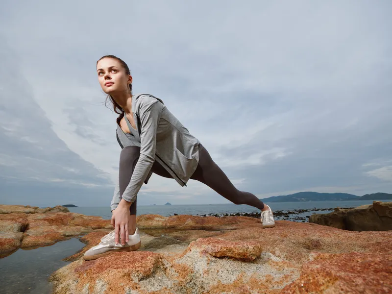 Young athletic woman stretching on rocky beach during cloudy day, showcasing dedication to fitness and healthy lifestyle with nature background