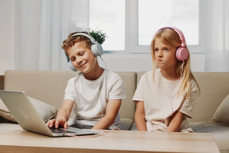 Two children wearing headphones using laptop on sofa, enjoying music and entertainment at home in bright living room.