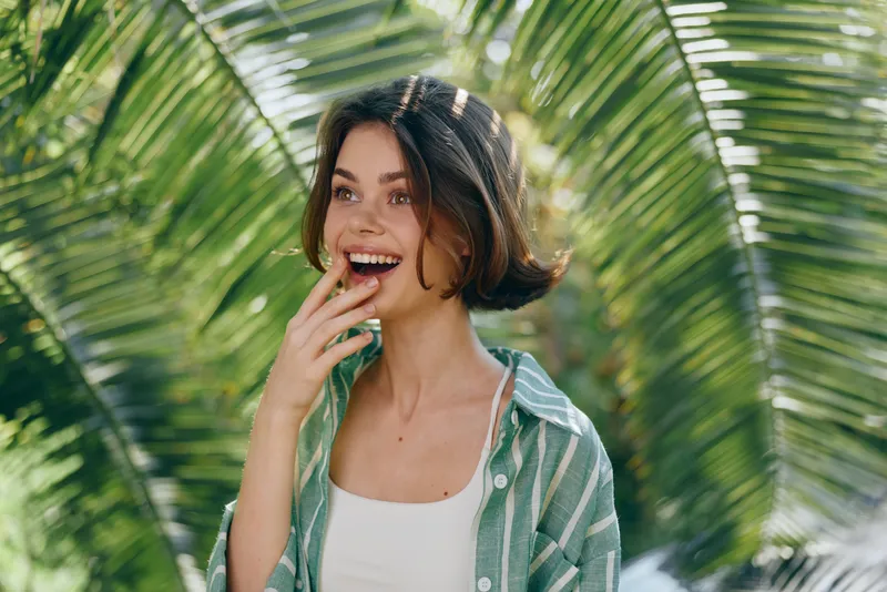 A cheerful young woman stands among lush palm leaves, smiling with a surprised expression.