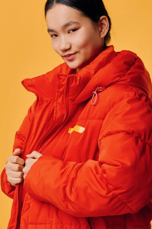Young woman wearing bright orange puffer jacket against yellow background, portrait with playful expression. Casual winter clothing, warm outerwear and confident pose in studio