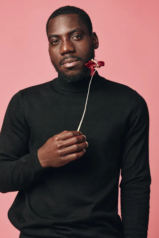 African man with dark skin wearing black turtleneck holding dried flower on pink background in creative conceptual visual style for portrait and fashion photography.
