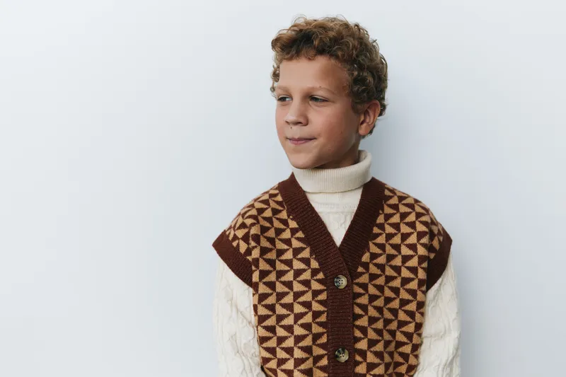Portrait of a young boy wearing a brown geometric knit vest over a cream turtleneck, standing against a pale wall. Soft light highlights texture and timeless vintage style.