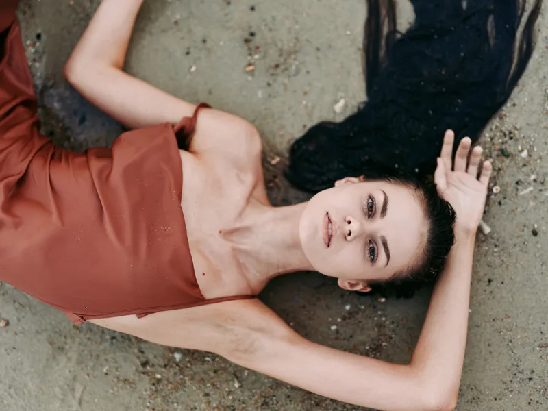 A woman lies on sandy ground in a rust red dress, gazing upward with a calm, introspective expression, creating a striking cinematic portrait against textured sand.