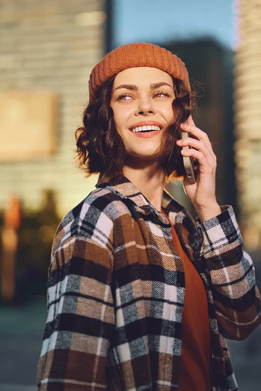 Woman on phone with a bright smile in urban street, candid beanie portrait at golden hour glow, authenticity, candid lifestyle and mindful living captured with emotional storytelling and modern