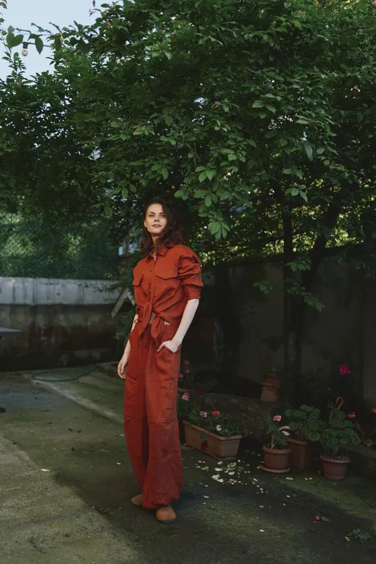 Woman in orange jumpsuit stands in a garden, relaxed pose under trees, natural light highlights her features, colorful pots and greenery create a calm outdoor lifestyle portrait