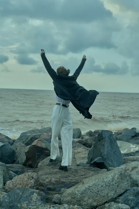 Man in a dark coat raises arms in a triumphant pose on a rocky coast, facing the sea under dramatic clouds, symbolizing celebration and outdoor resilience.