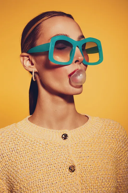 Young woman with large turquoise sunglasses blows a bubblegum bubble. She wears a yellow knitted sweater and gold hoop earrings against a bright yellow background. Fashion and fun concept.