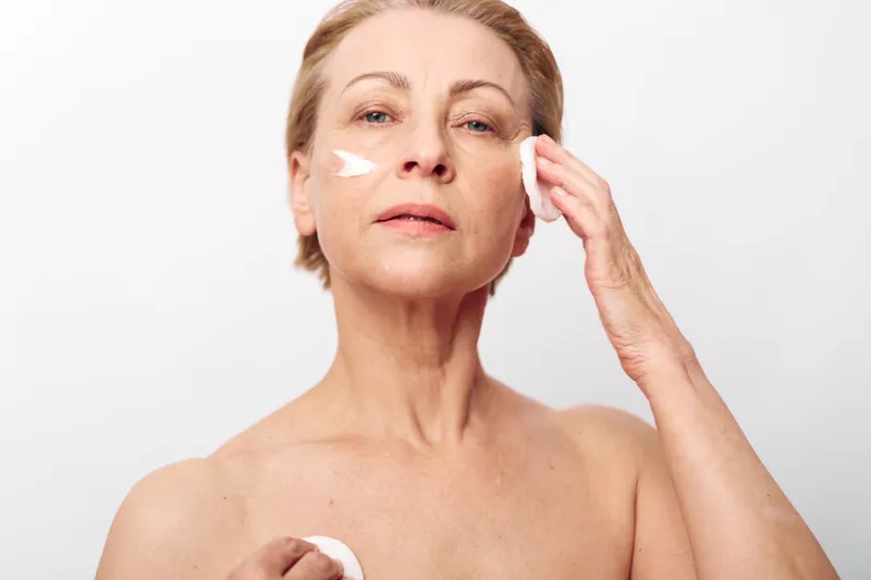 Mature woman applies face cream on cheek with cotton pad, showing skincare routine and self-care in bright studio. Concept of aging skin, beauty, and wellness.