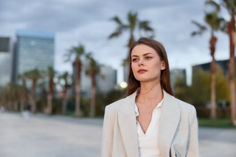 Young woman in light blazer expressing thoughtful calmness outdoors with urban background and palm trees, casual business style in soft natural lighting