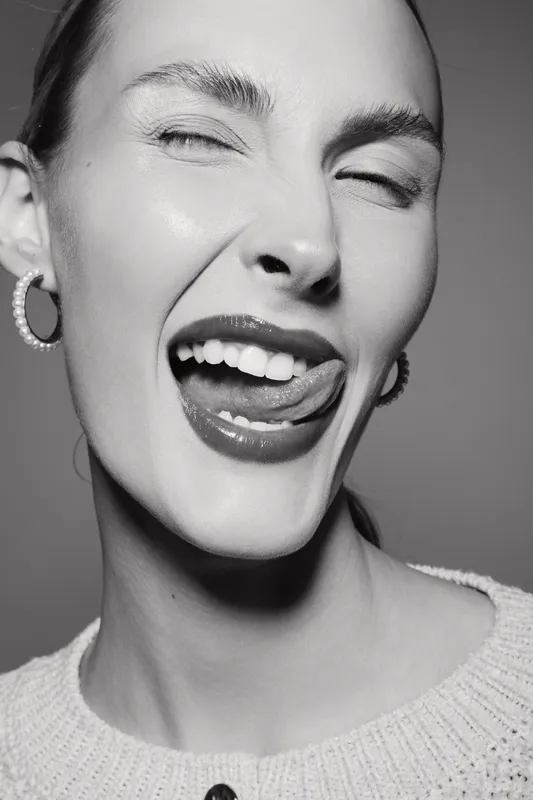 Close-up of a young white woman with light skin, smiling and winking with tongue out, wearing pearl earrings and knit sweater in black and white photo.