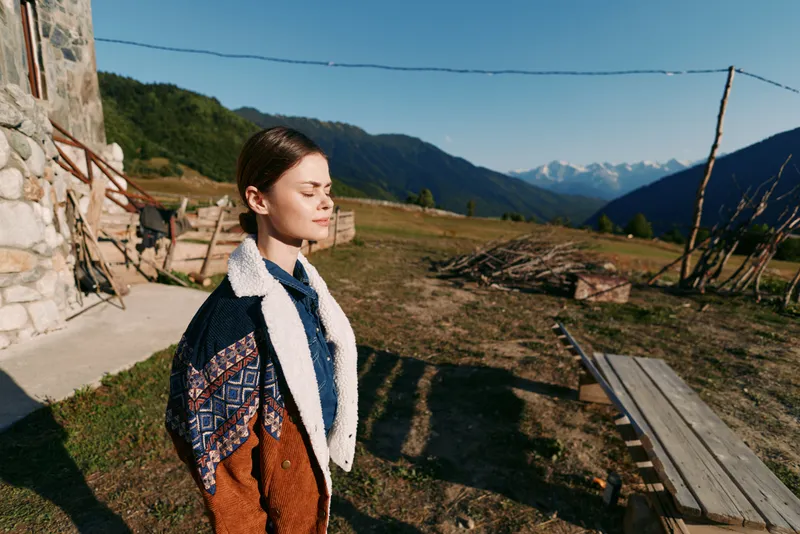 Woman portrait in sunlight by rustic cabin with closed eyes, standing in nature overlooking mountains and meadow. Serene outdoor scene with jacket, calm expression and peaceful landscape.