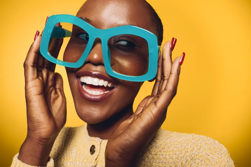 Smiling dark skinned African woman wearing oversized blue glasses poses against yellow background. Concept of fashion, joy, beauty, and vibrant lifestyle with playful accessories.