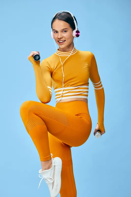 Fitness woman in bold orange athletic wear lifts a dumbbell in a dynamic workout scene; sport energy at the gym with headphones and confident pose.