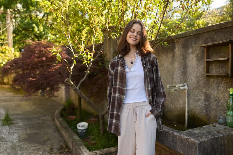 Smiling woman in a plaid shirt and light pants stands in a sunlit garden, showcasing a relaxed lifestyle and casual fashion in natural outdoor light