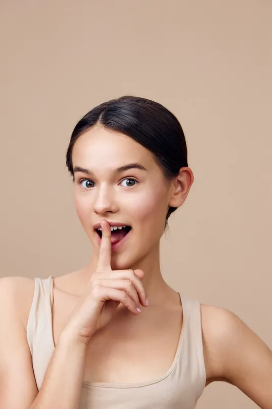 Young woman with a playful expression holding her finger to her lips, wearing a nude tank top, against a soft beige background, perfect for beauty and lifestyle concepts