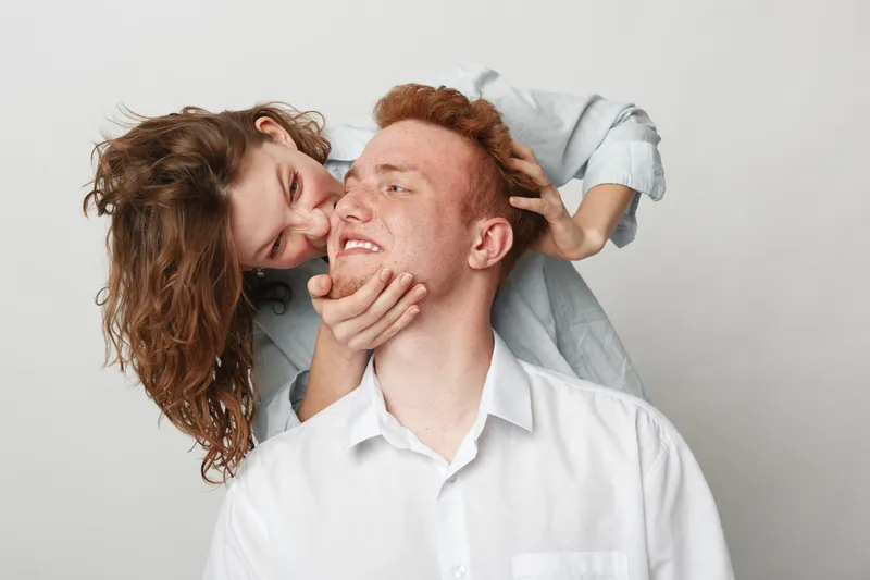 Playful young woman with brown hair teasing smiling redhead man in white shirt on light background. Concept of fun, relationship, happiness, and casual lifestyle together.