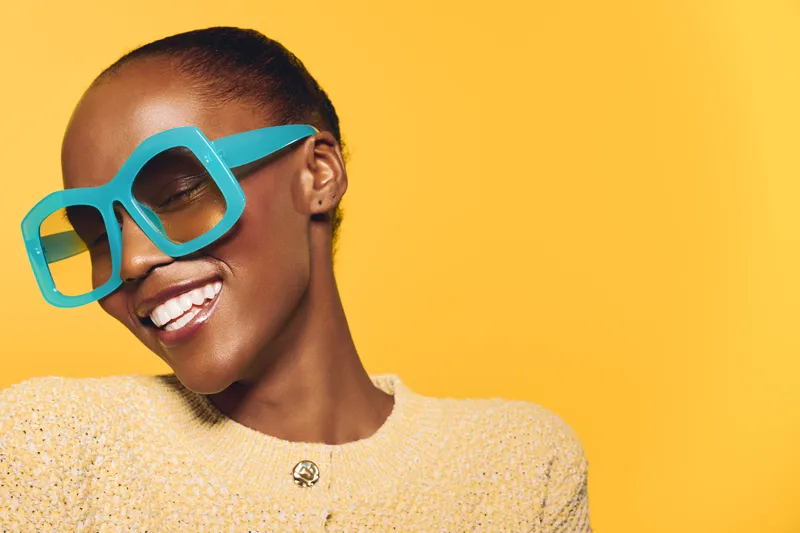 Smiling dark skinned African woman with short hair wears large blue sunglasses and yellow sweater. She poses happily against a bright yellow background. Concept of joy and fashion.