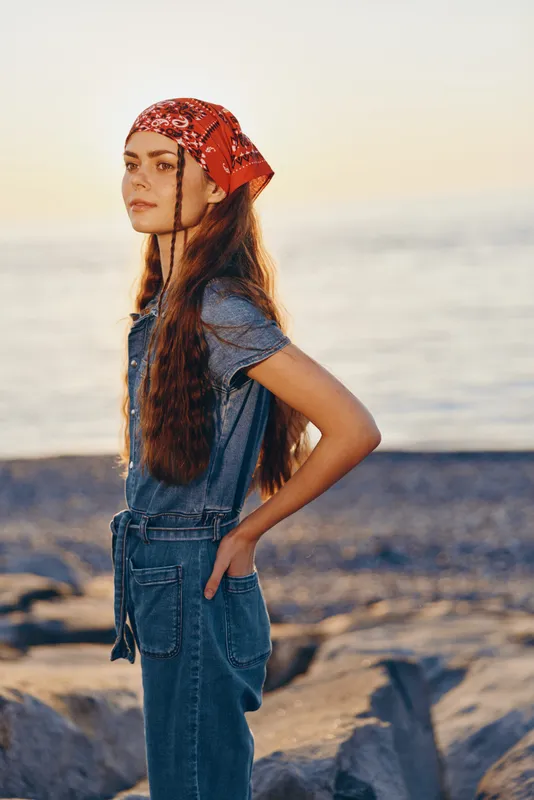 young woman wearing red bandana and denim overalls standing by the rocky seaside with calm expression during sunset golden hour